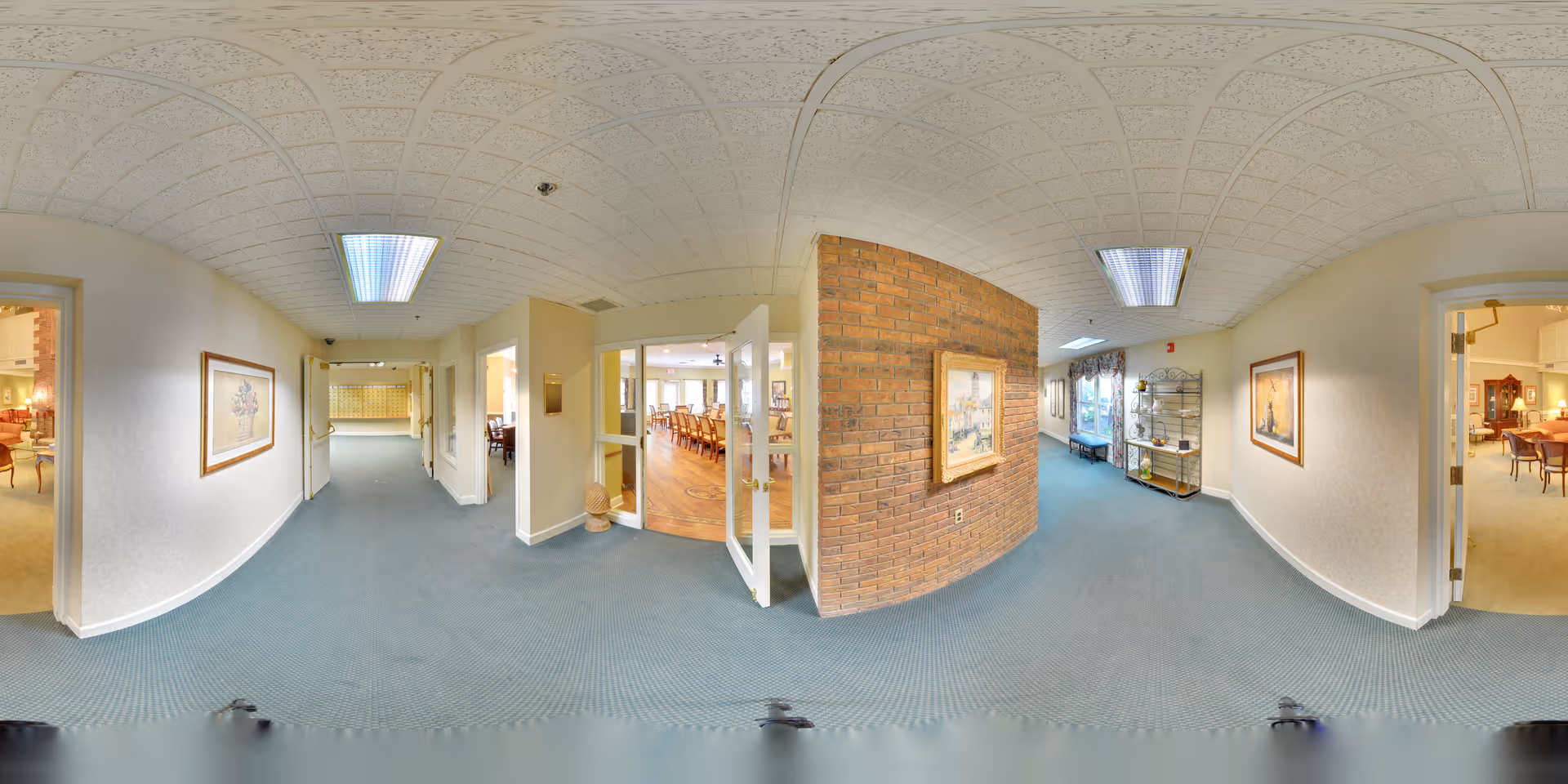 A wide hallway in a senior living facility with blue carpet and white walls. The hallway has framed artwork on the walls and leads to multiple rooms, including a dining area with wooden floors and tables visible through glass doors on the right. There is a brick accent wall with a painting, and the hallway is lit by ceiling lights.