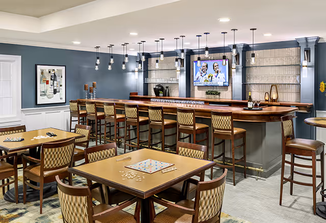 A well-lit senior living facility common area featuring a long wooden bar with high chairs, pendant lights hanging above, and a flat-screen TV mounted on the wall. In the foreground, there are tables with chairs arranged around them, one table set up with a Scrabble game. The walls are painted blue with white wainscoting, and there is framed artwork on one wall.