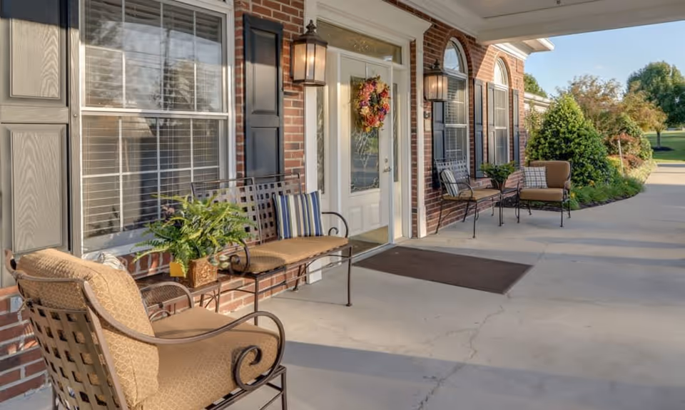 Front entrance patio with brick facade, a white door with wreath, outdoor seating, and potted plants along a concrete walkway.