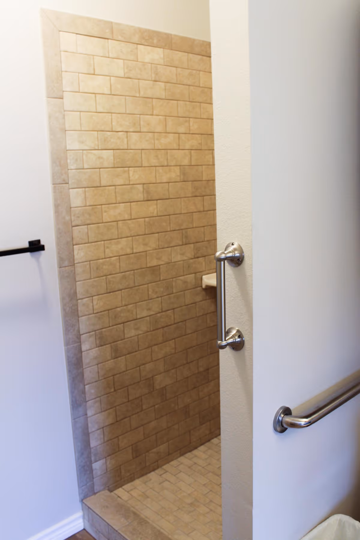 A tiled walk-in shower with beige rectangular tiles on the walls and smaller square tiles on the floor. There are two stainless steel grab bars mounted on the walls near the shower entrance, one vertical and one horizontal, indicating accessibility features.