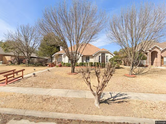 Front exterior view of a single-story residential building with a light-colored brick facade and a brown shingle roof. The yard has dry grass, several leafless trees, a concrete sidewalk, and a small wooden bridge on the left side. The sky is clear and blue.