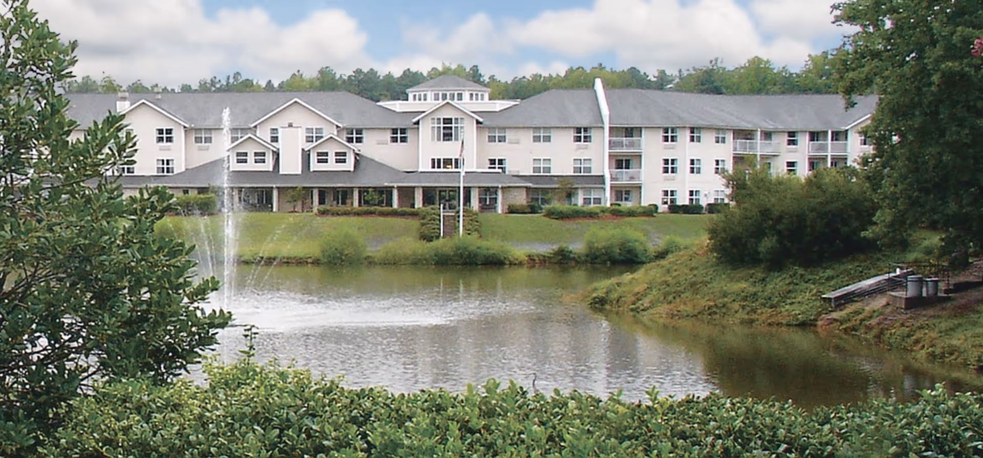 A large multi-story senior living facility building with white exterior walls and a gray roof, situated behind a pond with a water fountain. The surrounding area includes green bushes, trees, and a grassy landscape under a partly cloudy sky.