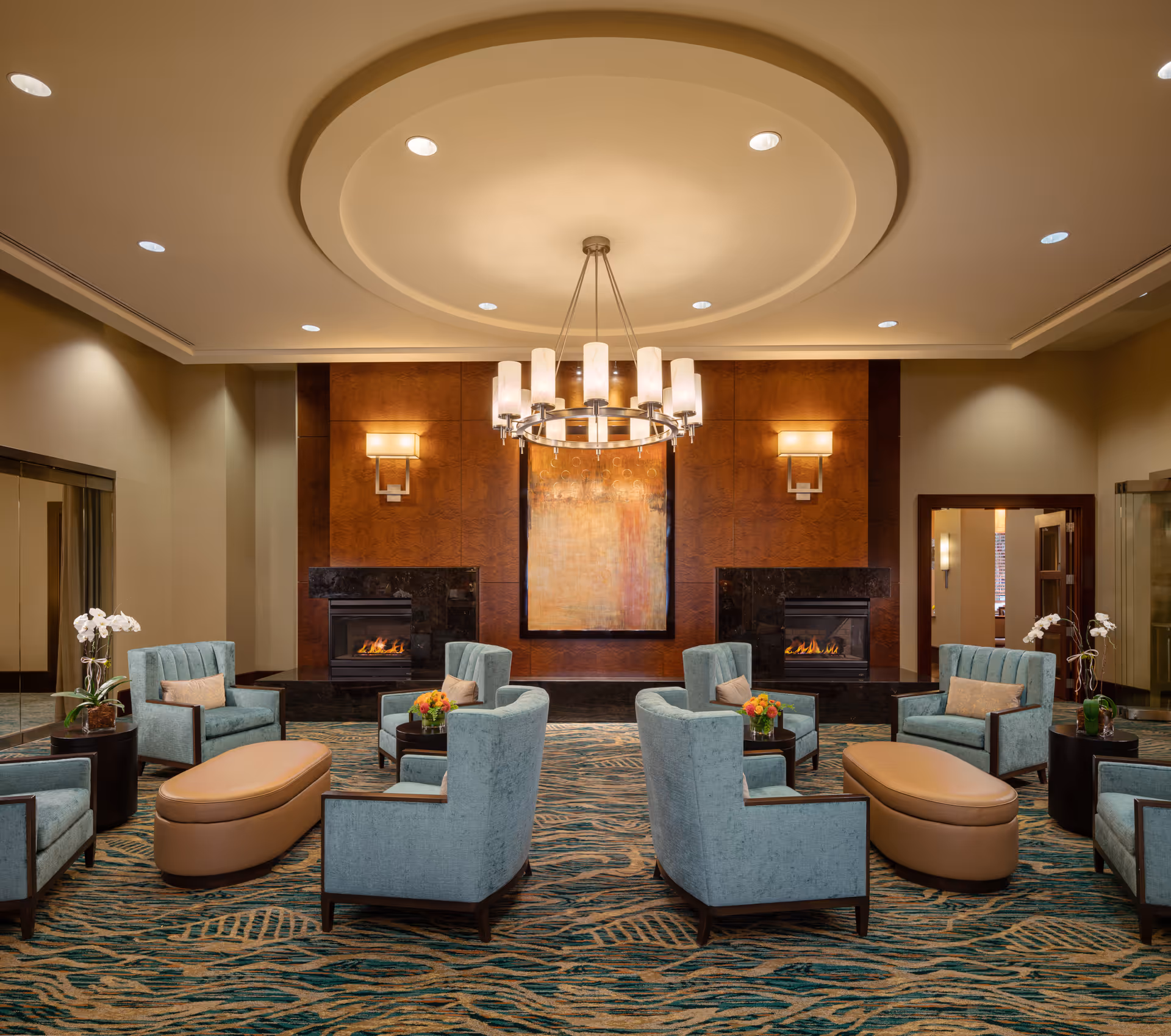 A spacious and elegant living room area with a circular chandelier hanging from a recessed ceiling. The room features two modern fireplaces set into a wooden accent wall with a large abstract painting in the center. There are six light blue upholstered armchairs arranged around two oval tan ottomans and small tables with flower arrangements. The carpet has a blue and beige pattern, and there are white orchids on side tables near the walls.