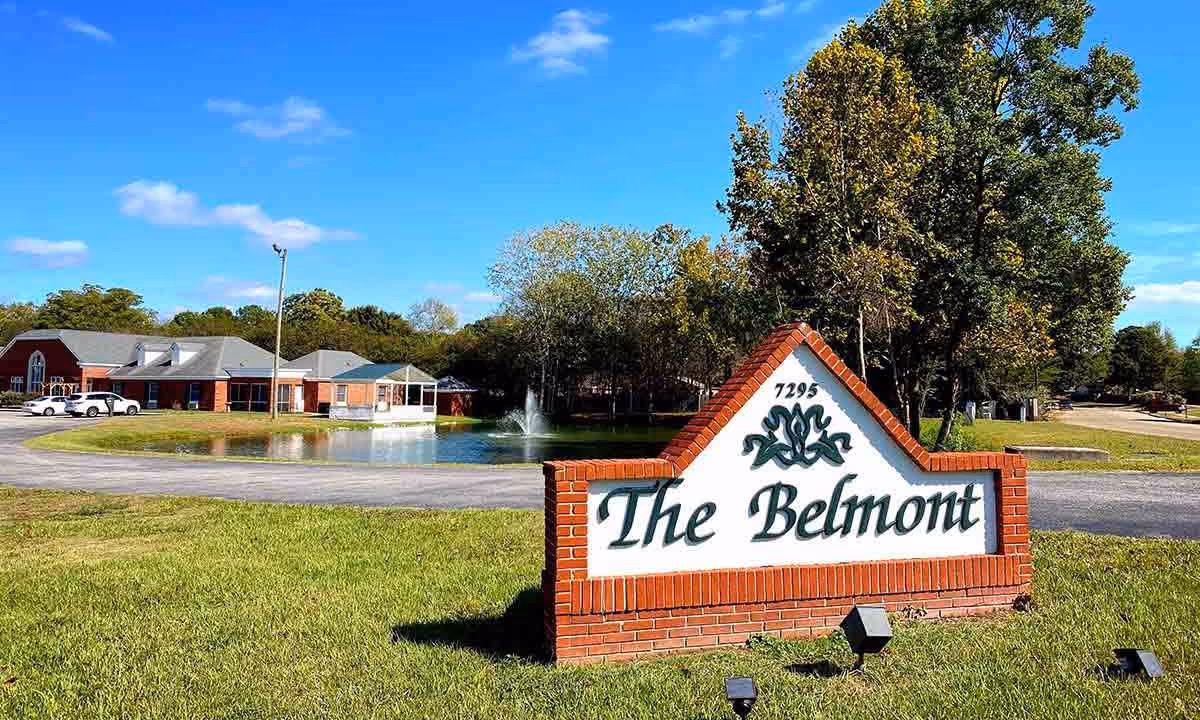 Exterior view of The Belmont assisted living facility with a large brick sign in the foreground displaying the name and address. The building is visible in the background with a pond and fountain surrounded by trees and a clear blue sky.