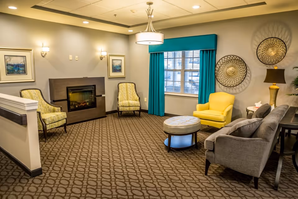 A cozy living room area with patterned carpet, a modern electric fireplace, two yellow patterned armchairs, a bright yellow armchair, a gray sofa, a round ottoman, and a window with teal curtains. The walls are decorated with two framed paintings and two circular metal wall art pieces. A table lamp is placed on a side table next to the sofa.