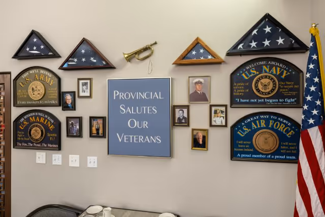 Wall display honoring veterans with framed photos, folded American flags in triangular cases, military plaques for U.S. Army, U.S. Marine Corps, U.S. Navy, and U.S. Air Force, a small brass bugle, and a sign reading 'Provincial Salutes Our Veterans'. An American flag stands to the right side of the display.