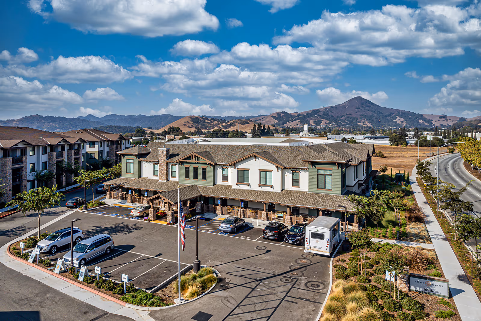 Aerial view of Loma Clara Senior Living facility showing a two-story building with a parking lot in front, several parked cars, an American flag on a flagpole, landscaped greenery, and a mountainous background under a partly cloudy blue sky.
