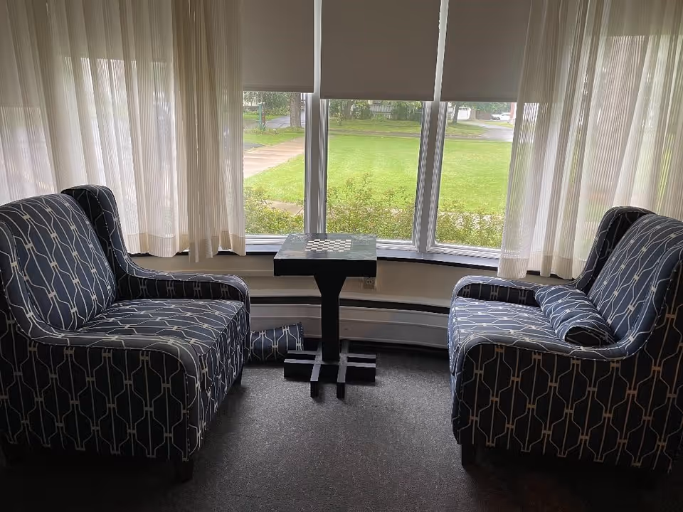Two patterned armchairs facing each other with a small black table with a chessboard pattern between them, positioned in front of a large window with sheer curtains and a view of green grass and a sidewalk outside.