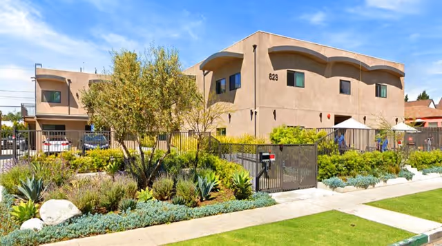 Exterior view of a two-story beige building with the number 823 on the wall, surrounded by a black metal fence and landscaped garden with various plants and shrubs under a blue sky.