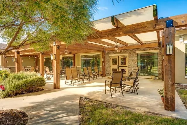 Covered outdoor patio with a wooden pergola, multiple metal chairs and tables, and surrounding landscaping.