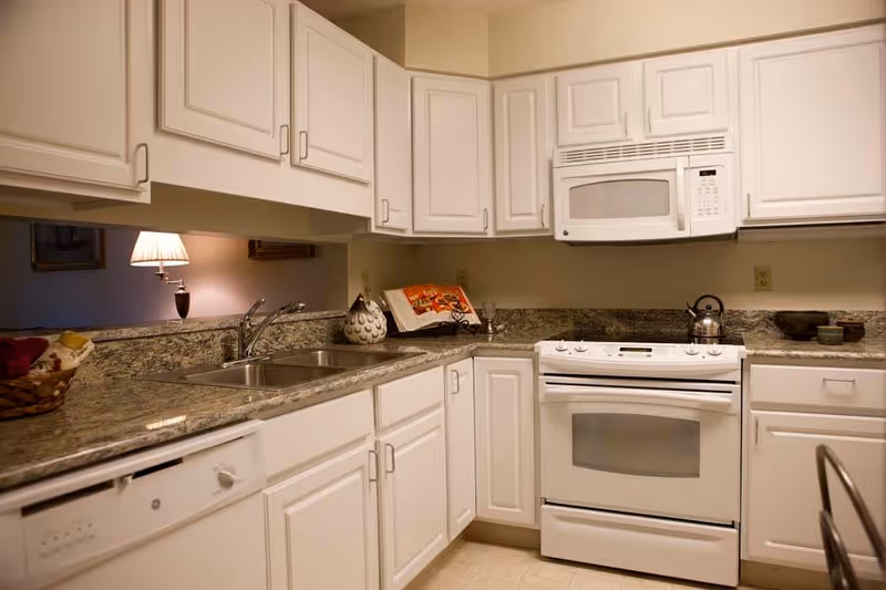 A clean kitchen with white cabinets and granite countertops. The kitchen features a double sink, a white dishwasher, a white stove with an oven, and a white microwave above the stove. There is a kettle on the stove, a decorative vase, a cookbook on a stand, and a basket with cloth on the countertop. A lamp and framed pictures are visible in the adjacent room through a pass-through window.