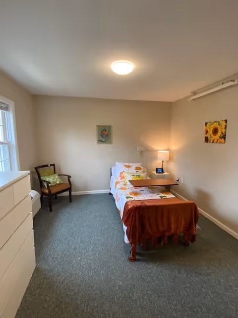 A simple, clean bedroom with a single bed covered in a floral bedspread and a rust-colored throw blanket. There is a wooden chair with a green cushion near a window, a white dresser, a bedside table with a lamp, and two framed pictures on the beige walls. The room has a carpeted floor and soft lighting.