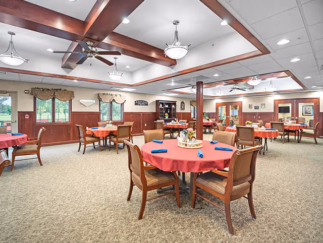 A spacious dining room with multiple round tables covered with red tablecloths and set with blue napkins. Each table has a centerpiece with flowers. The room features wooden beams on the ceiling, ceiling fans, and large windows with valances allowing natural light to enter. There are wooden chairs around each table and a carpeted floor.