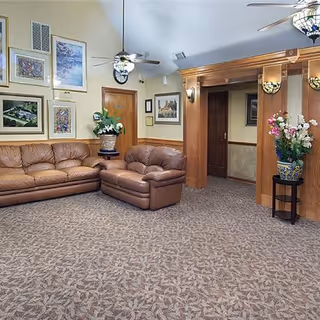 A cozy living room area in an assisted living facility featuring two brown leather sofas, patterned carpet, several framed pictures on the walls, ceiling fans with lights, and decorative plants in pots placed on stands.
