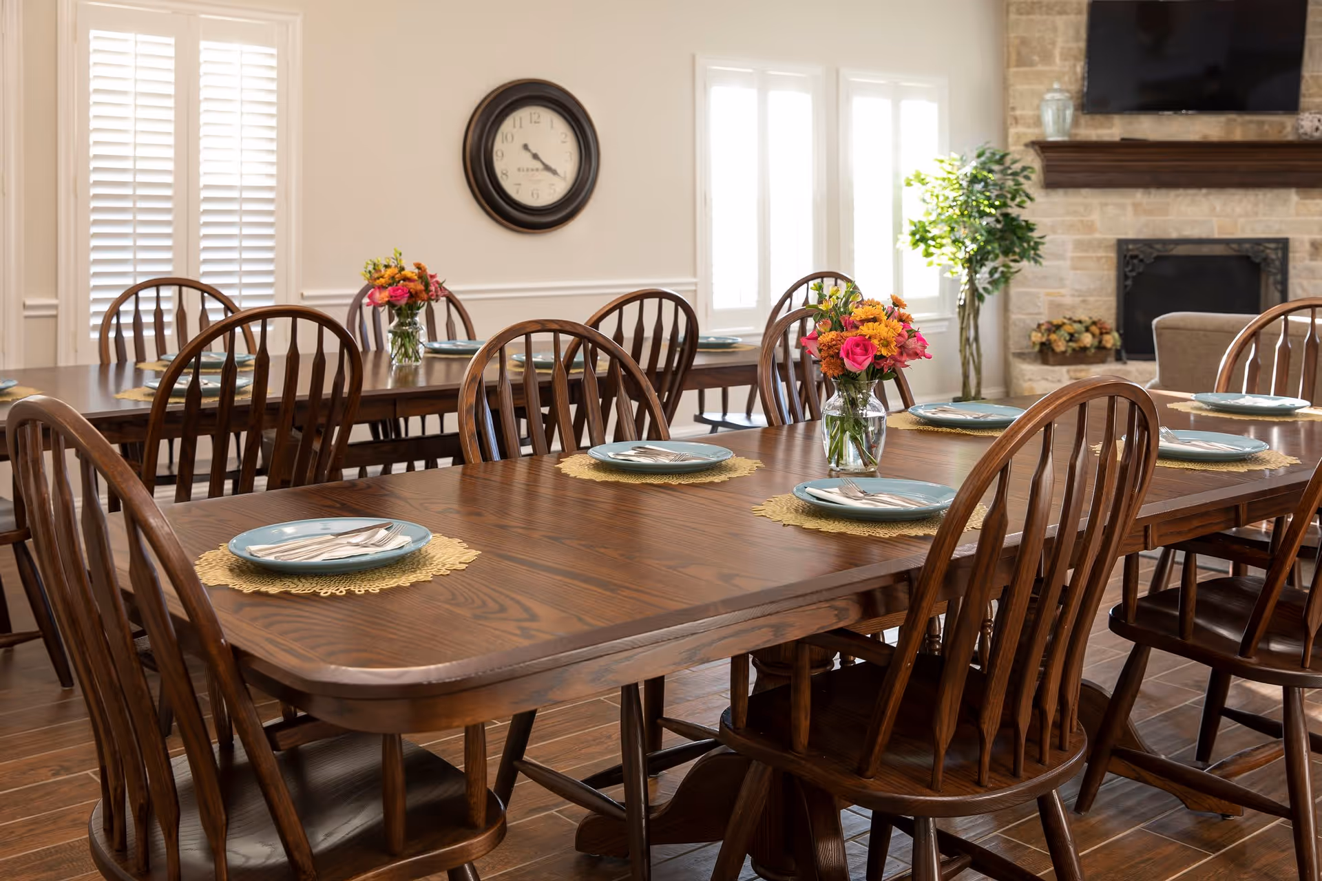 A dining room with two wooden tables set with blue plates and silverware on yellow placemats. Each table has a vase with colorful flowers. The room features wooden chairs, a wall clock, large windows with white shutters, a stone fireplace, and a potted plant.