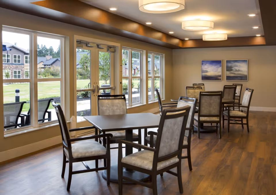 A bright dining area with several square tables and cushioned chairs arranged neatly. Large windows and glass doors provide a view of a green outdoor space with buildings in the background. The room features wooden flooring, recessed lighting, and two framed landscape paintings on the far wall.
