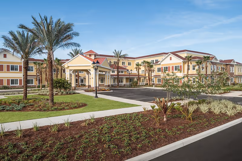 Exterior view of Buffalo Crossings Assisted Living facility showing a large, multi-story building with yellow and beige walls, red roof accents, palm trees, landscaped garden beds, and a paved parking area under a clear blue sky.