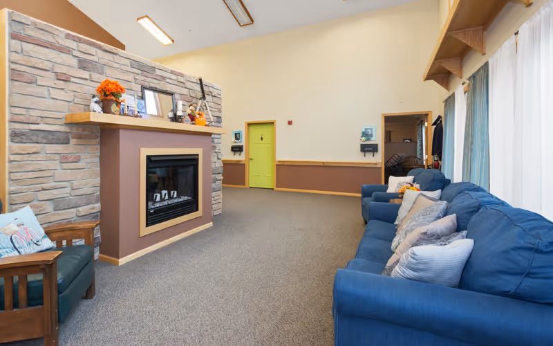 Bright assisted-living common room with a stone-faced fireplace, blue sofas along large windows, and a lime-green door at the back.