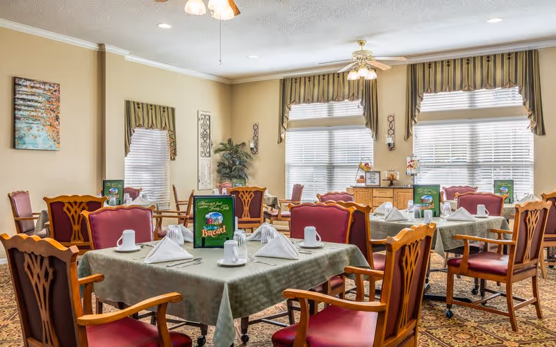 A bright dining room in a senior living facility with multiple tables covered in green tablecloths, each set with white napkins, cups, and silverware. The chairs have wooden frames with red cushioned seats and backs. Large windows with striped valances allow natural light to fill the room. There is a decorative plant in the corner and framed artwork on the walls.