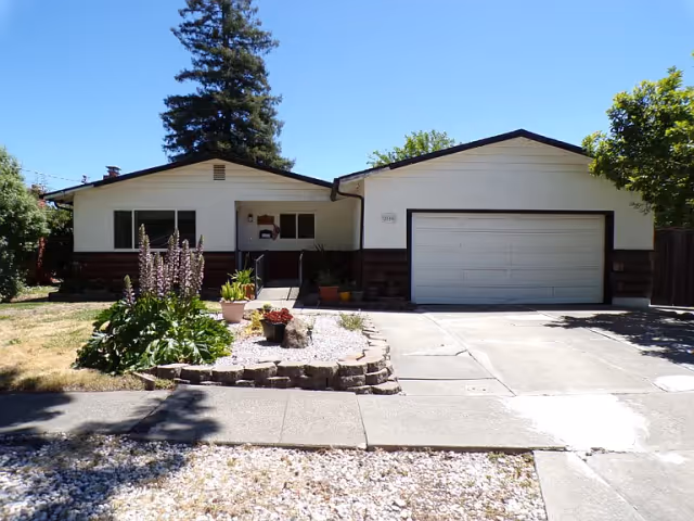 Single-story house with an attached two-car garage, front yard landscaping, and a concrete driveway under a clear blue sky.