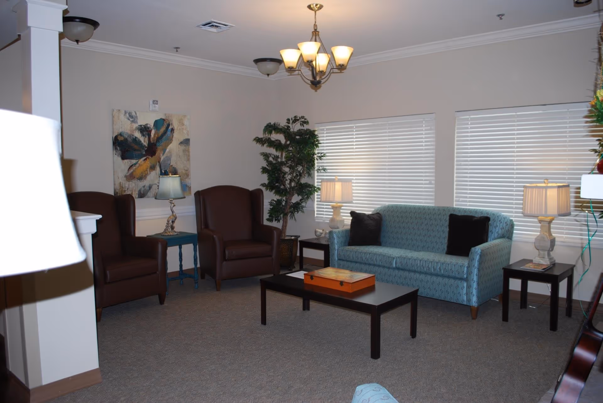 A cozy living room with two brown armchairs, a blue patterned sofa with two black cushions, a black coffee table with a wooden box on it, two side tables with lamps, a large potted plant, and a painting of a flower on the wall. The room has beige walls, carpeted floor, and white blinds covering the windows.