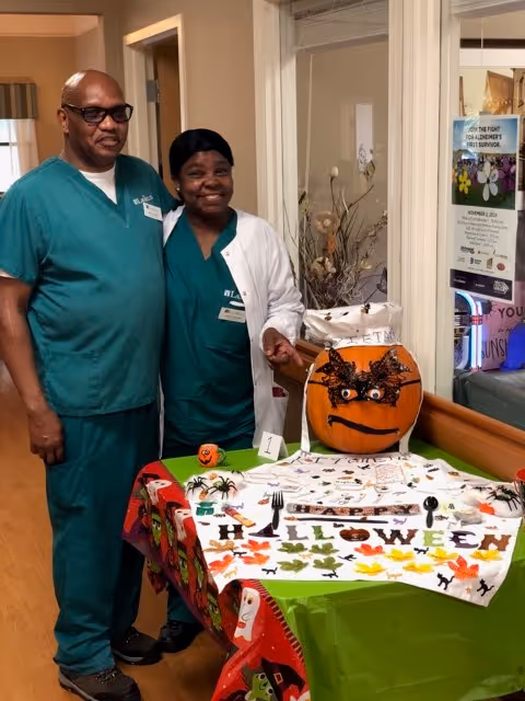 Two healthcare workers in green scrubs and white coat standing next to a table decorated with Halloween-themed items including a pumpkin with a spider mask, small spider decorations, and a tablecloth with Halloween motifs in a hallway of a senior living facility.