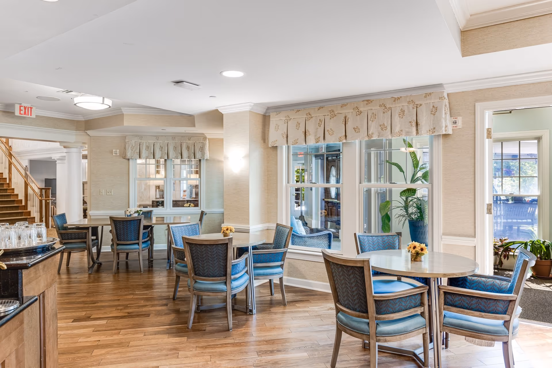Well-lit dining area with round tables and upholstered chairs on hardwood floors, large windows and potted plants.