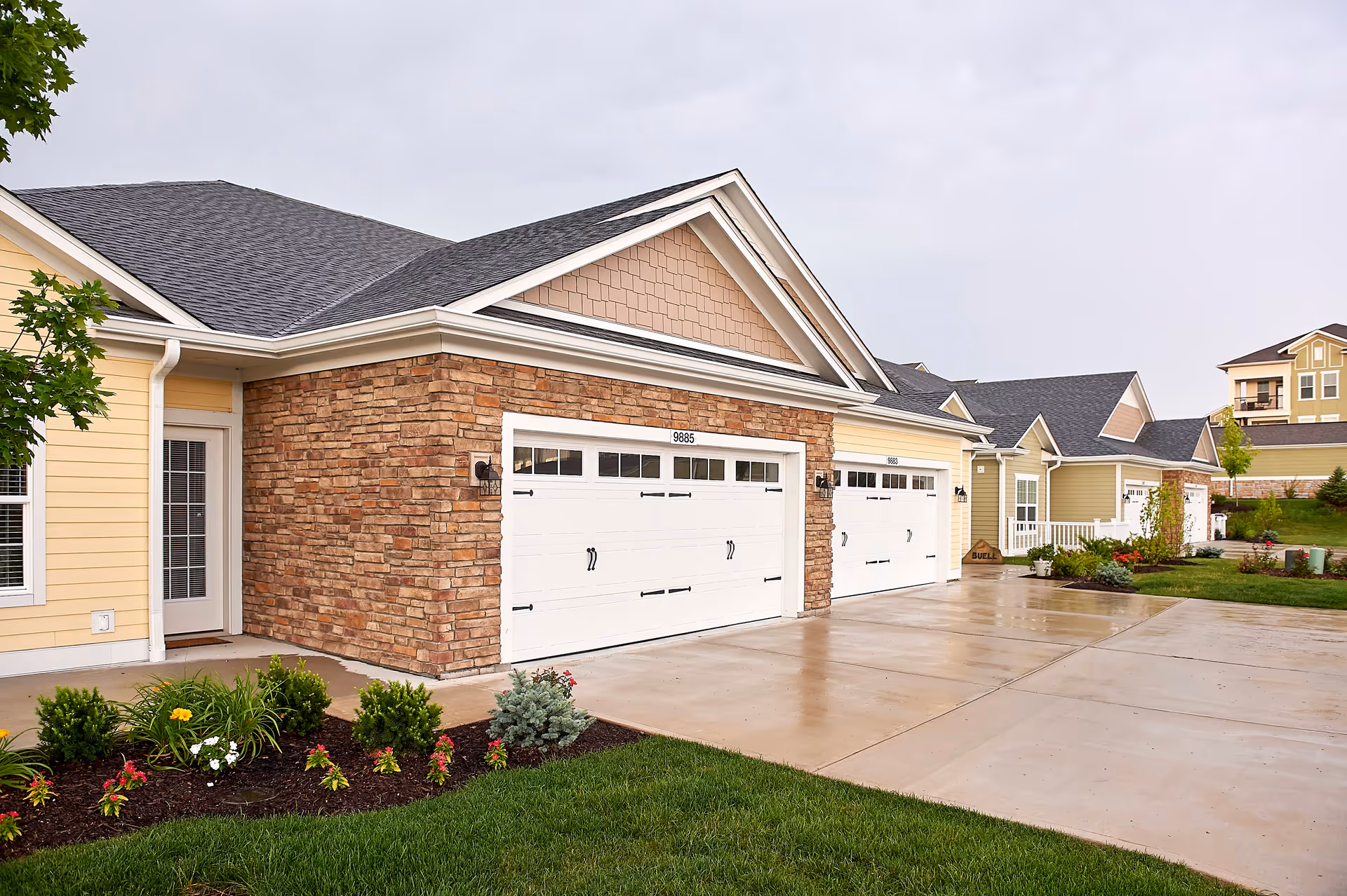 Exterior view of a residential building with beige siding and stone accents, featuring two white garage doors and a concrete driveway. There are small landscaped garden beds with green shrubs and flowers in front of the building, and other similar buildings are visible in the background under a cloudy sky.