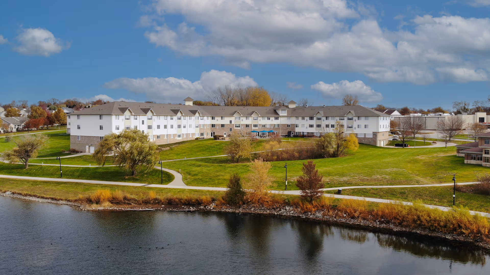 Large multi-story senior living building beside a lake with green lawns, walking paths, and trees under a partly cloudy sky.