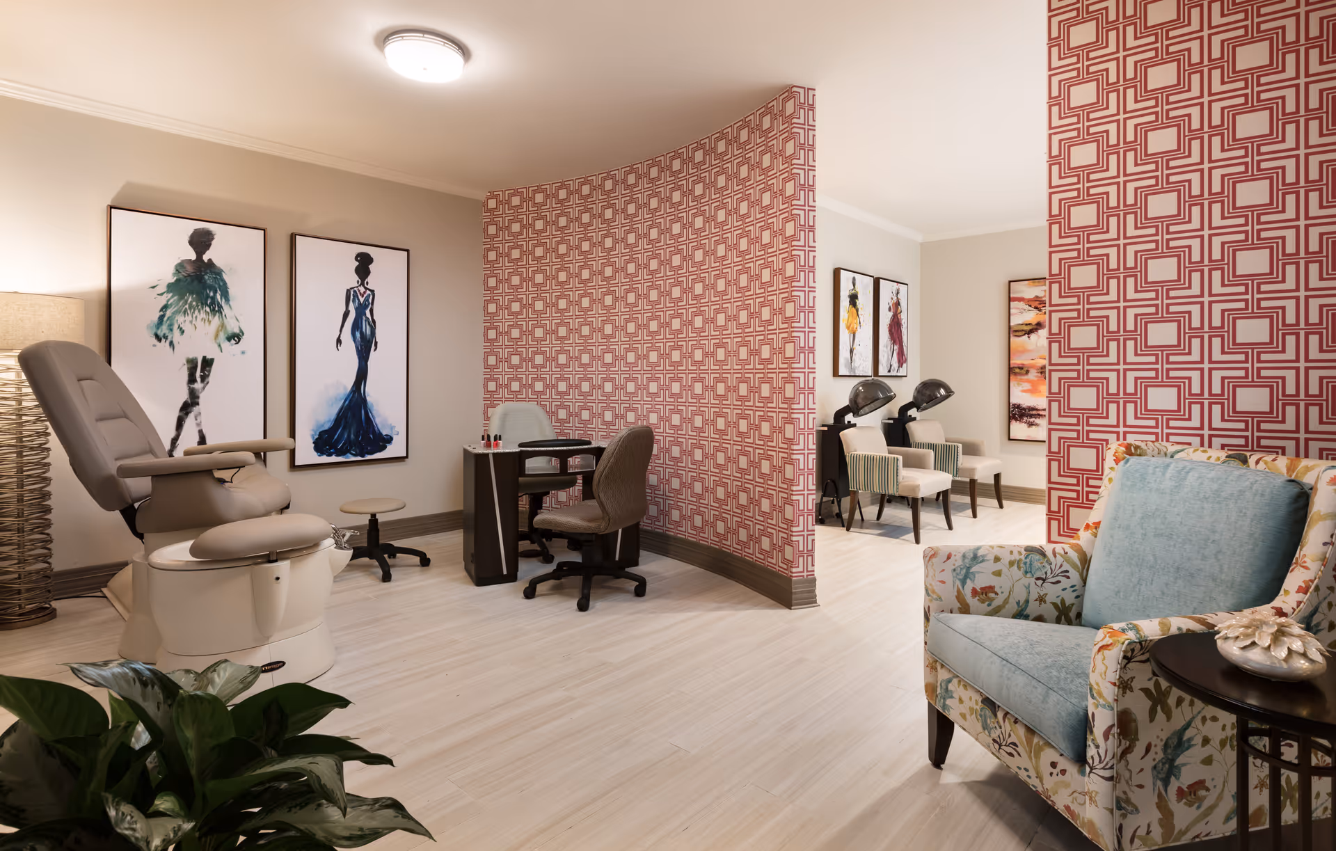 Interior view of a stylish salon area in a senior living facility with a pedicure chair, nail technician desk, two hair drying chairs, patterned red and white wallpaper, framed fashion artwork on the walls, a floral armchair with a blue cushion, and a potted plant in the foreground.