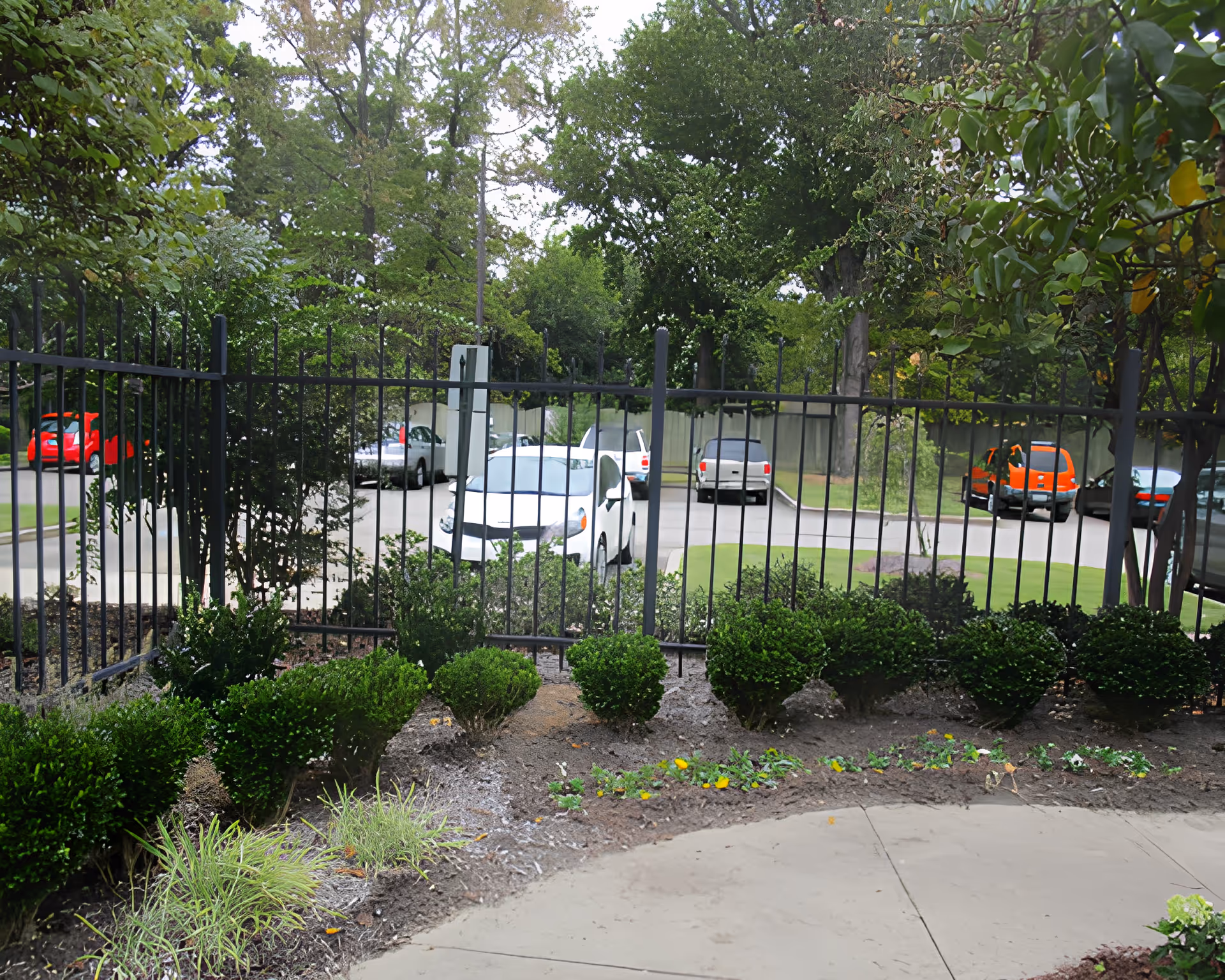 View of a parking lot with several cars behind a black metal fence, surrounded by green trees and bushes, with a curved concrete pathway and landscaped garden in the foreground.