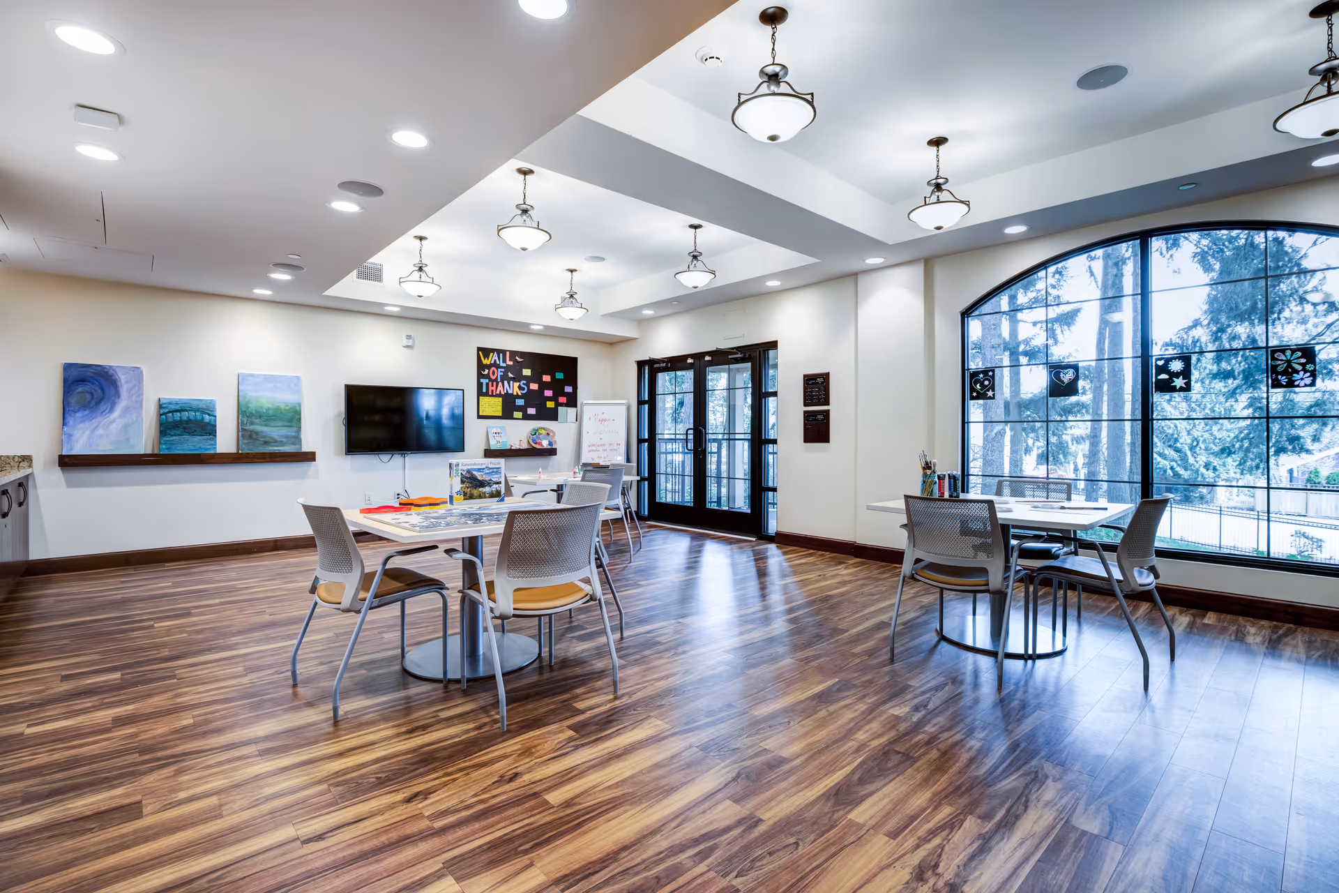 A spacious, well-lit room with wooden flooring featuring several tables and chairs arranged for activities. The room has a large arched window with a view of trees outside, a wall-mounted TV, colorful artwork on the walls, and a bulletin board labeled 'Wall of Thanks'. Ceiling lights and pendant lamps illuminate the space.