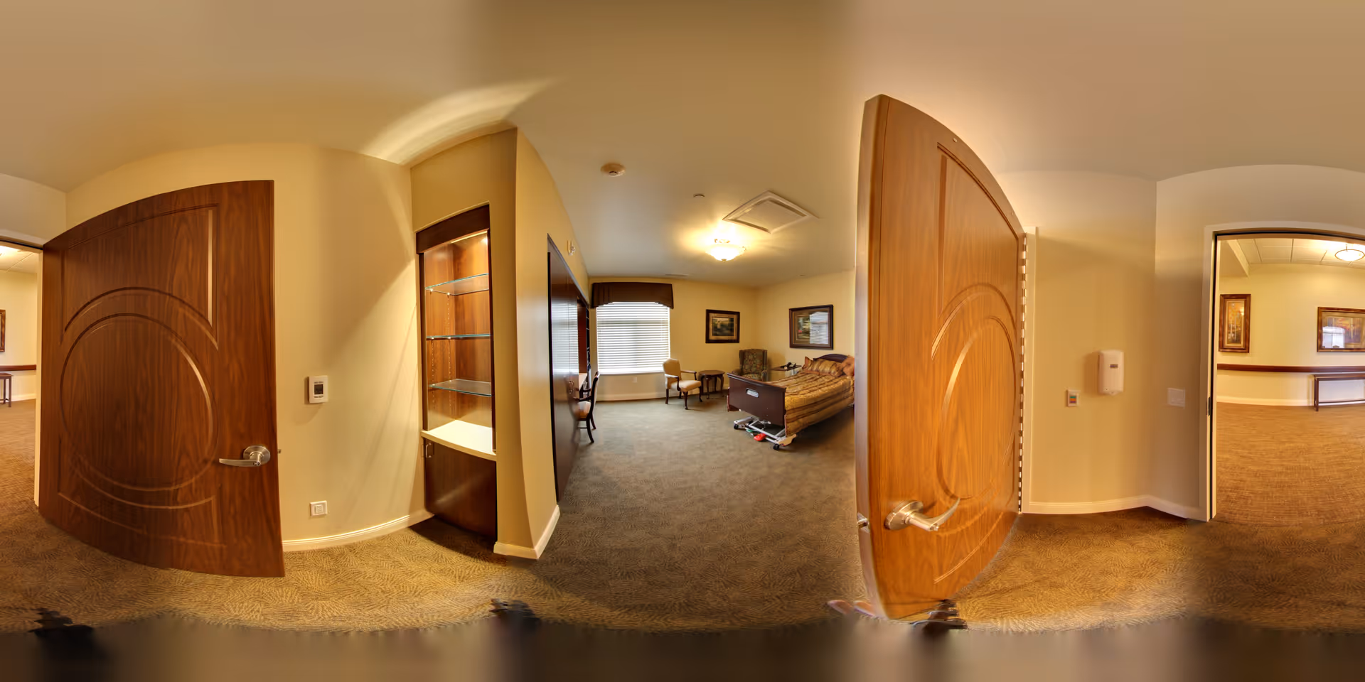 A spacious, well-lit bedroom in a senior living facility with beige walls and carpeted floor. The room features a hospital-style bed with side rails, a wooden headboard, and a blanket. There are two chairs and a small table near a large window with blinds. The room has wooden doors, one open showing a hallway with framed artwork on the walls. A built-in shelving unit is visible near the entrance.