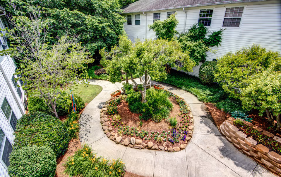 A landscaped outdoor courtyard with a circular flower bed in the center, surrounded by a concrete walkway. The courtyard is bordered by green bushes, trees, and a white building with multiple windows.