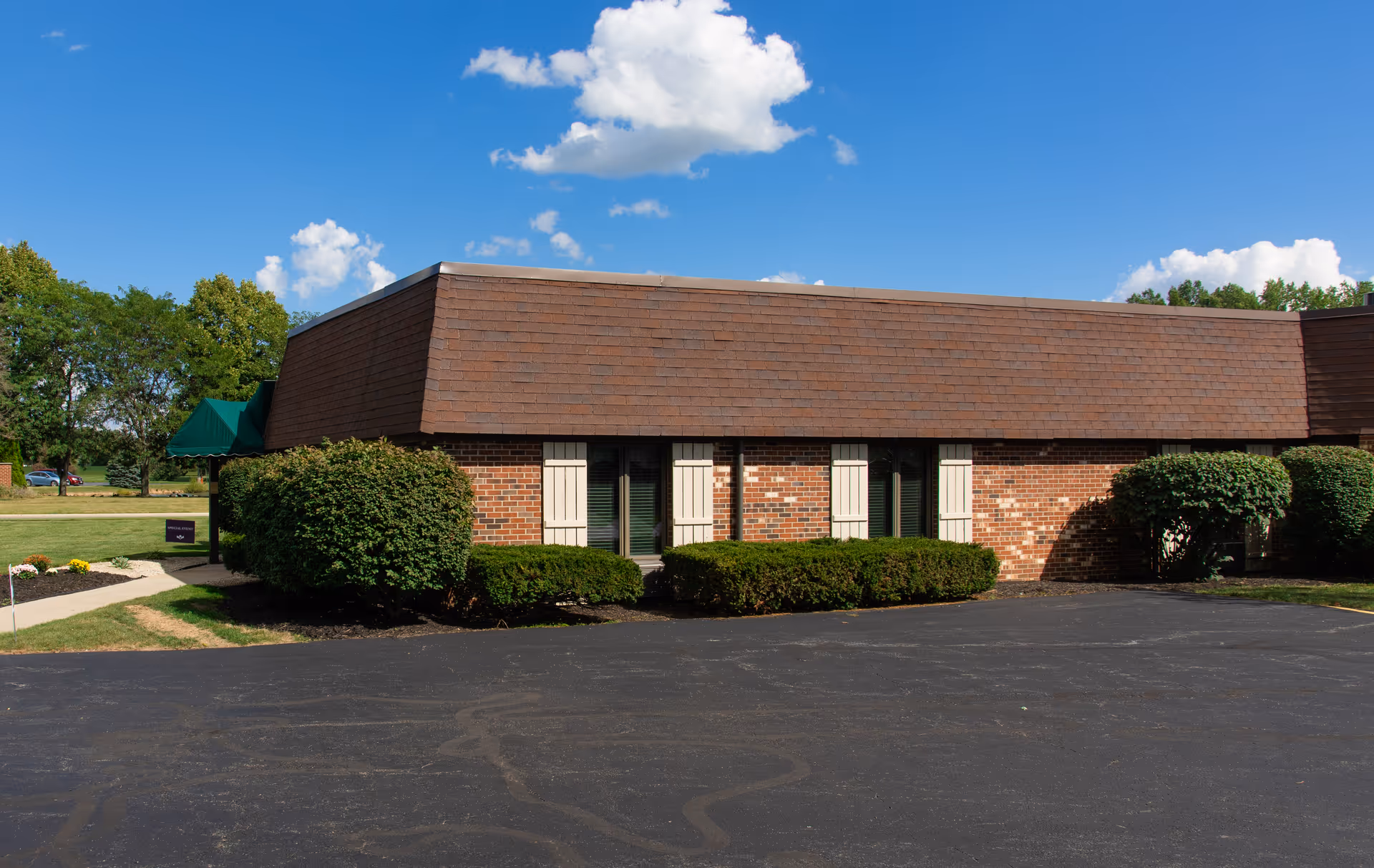 Exterior view of a single-story brick building with a brown shingled roof, surrounded by neatly trimmed bushes and a paved driveway under a bright blue sky with scattered clouds.