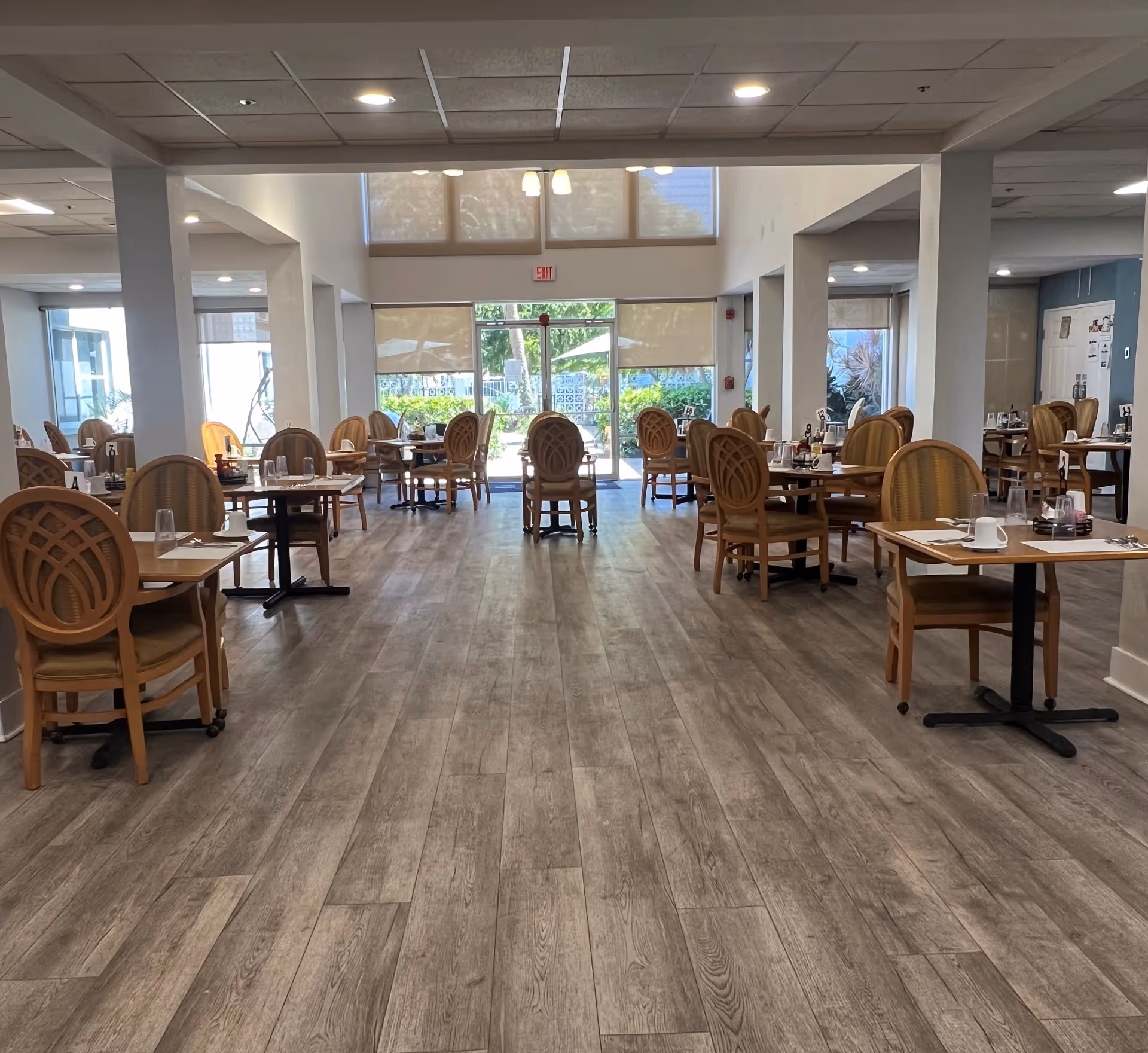 A spacious dining room with multiple wooden tables and chairs arranged neatly. Each table is set with placemats, cups, glasses, and utensils. Large windows and glass doors at the far end allow natural light to brighten the room, and the floor is covered with wood-patterned flooring.