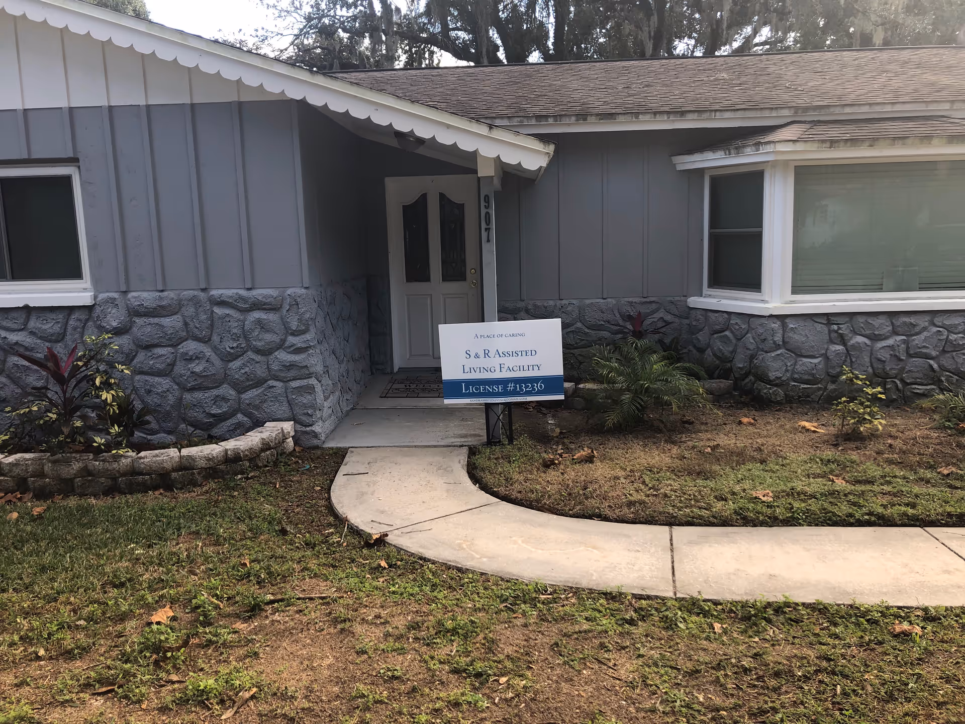 Exterior view of a single-story building with gray stone and siding walls, a white door with the number 907, a curved concrete walkway leading to the entrance, and a sign in front that reads 'S & R Assisted Living Facility License #13236'. There are small plants and a grassy area around the walkway.