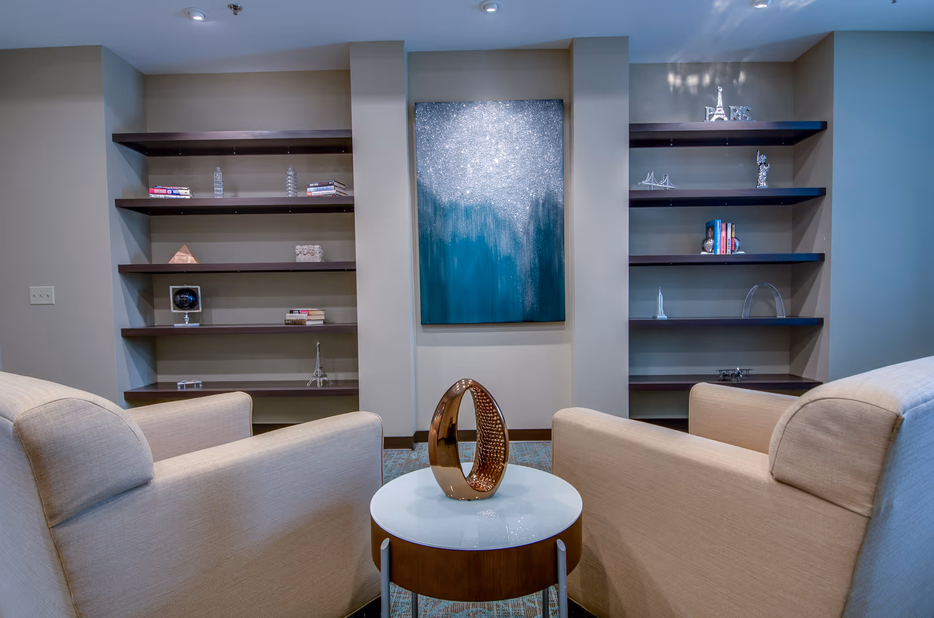 A cozy sitting area with two beige armchairs facing a small round table with a decorative sculpture. Behind the table is a wall with built-in dark wooden shelves holding books and small decorative items. A large abstract painting with blue and silver tones hangs in the center between the shelves.