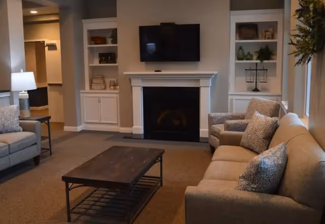 A cozy living room with a beige sofa and armchair, a wooden coffee table, a fireplace with a white mantel, built-in shelves on either side of the fireplace, and a flat-screen TV mounted above the fireplace. A lamp is on a side table next to the sofa, and a window with a wreath is visible on the right side.