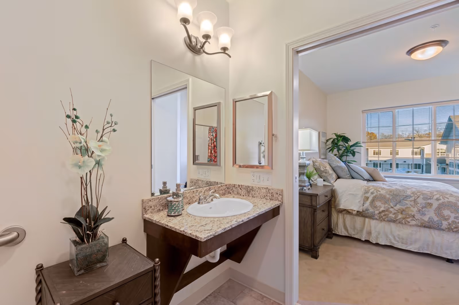 View of a bathroom sink with a granite countertop and a large mirror above it. To the left of the sink is a small wooden cabinet with a decorative plant on top. Through an open doorway, a bedroom is visible with a bed, nightstand, lamp, and a large window showing an exterior view of neighboring buildings.