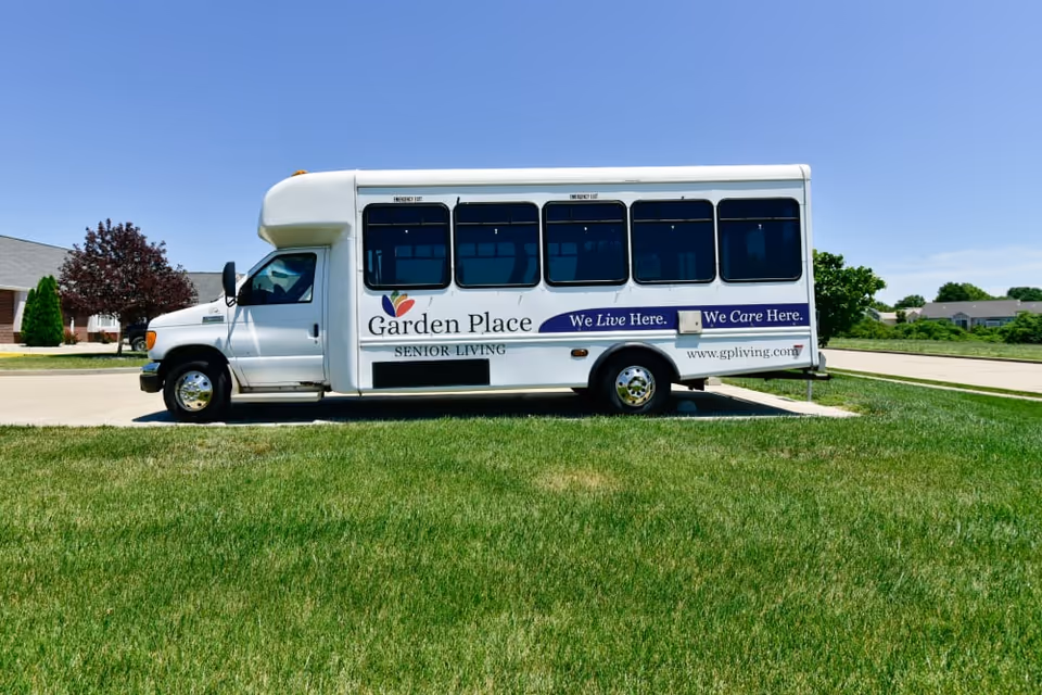 A white Garden Place Senior Living shuttle bus parked on a driveway beside a grassy lawn under a clear blue sky.