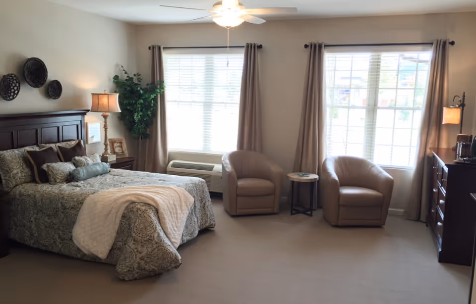 Spacious, sunlit bedroom with a made bed, bedside lamps, two beige armchairs by large windows, and wooden dressers.