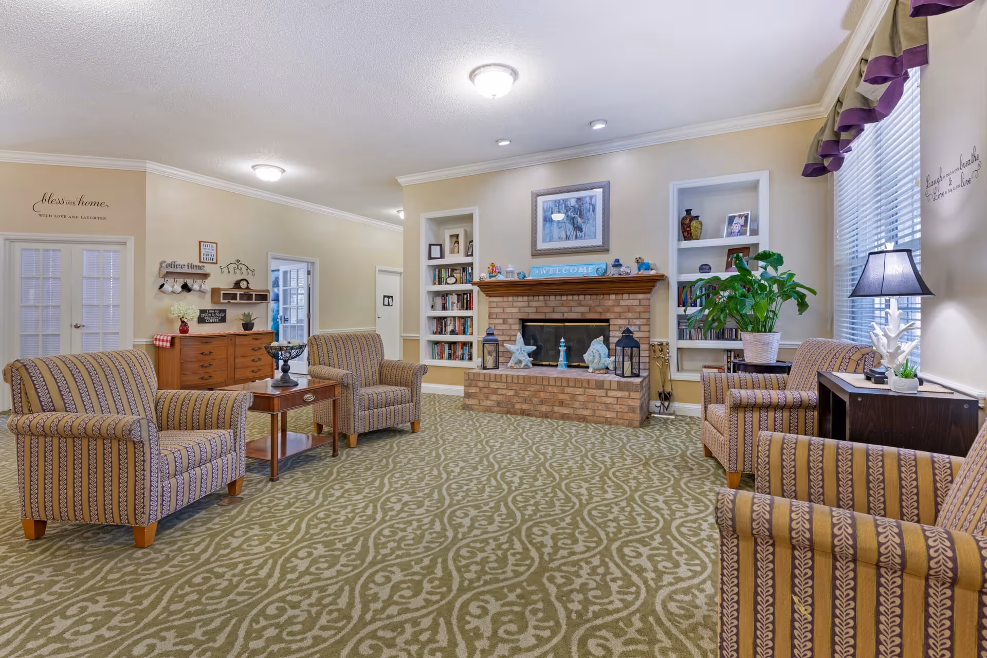 A cozy living room with patterned carpet and four striped armchairs arranged around a wooden coffee table. A brick fireplace with decorative items and a framed picture above it is centered on the far wall, flanked by built-in bookshelves filled with books and decor. A window with blinds and a valance is on the right, next to a side table with a lamp and a potted plant. The walls are painted beige with inspirational wall decals, and there is a wooden dresser with coffee mugs and signs on the left side of the room.