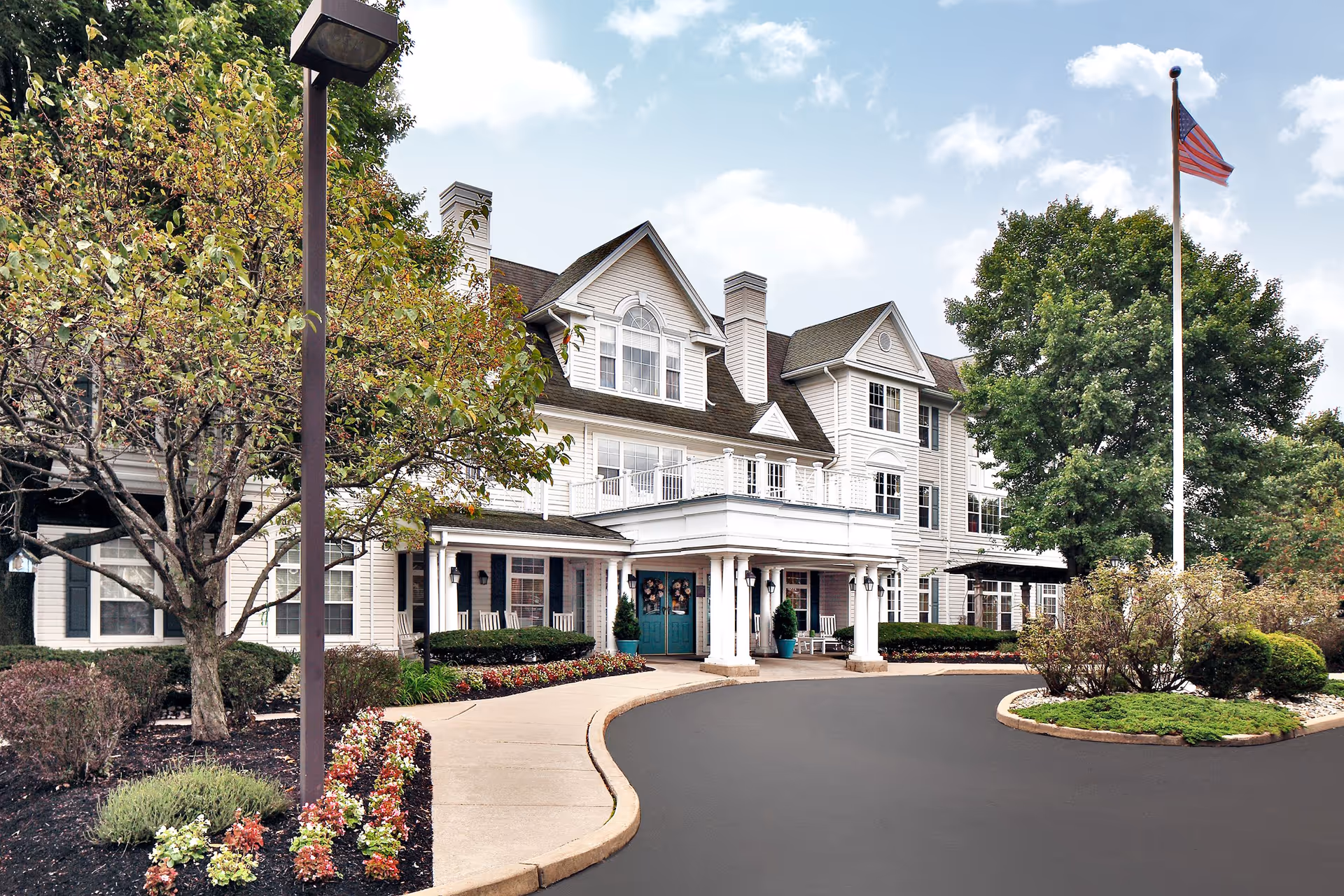 Exterior view of a senior living residence with curved driveway, landscaping, and flag.