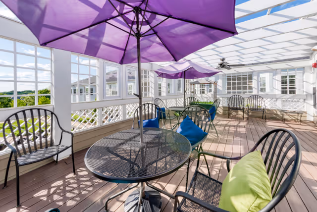 A bright outdoor patio area with black metal tables and chairs, some with colorful cushions in green and blue. Purple umbrellas provide shade, and the space is enclosed with white lattice and large windows. The floor is wooden, and the area is covered with a white pergola allowing sunlight to filter through.