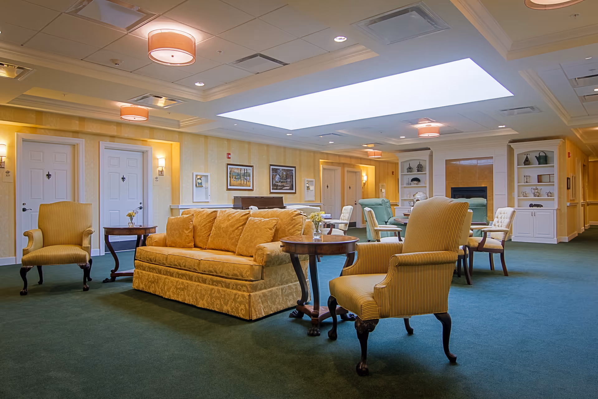 A spacious and well-lit common area in a senior living facility featuring a yellow upholstered sofa and matching armchairs arranged around wooden side tables. The room has green carpet flooring, soft yellow walls with framed artwork, and a large skylight in the ceiling. In the background, there are additional seating areas with armchairs and a fireplace flanked by built-in shelves.