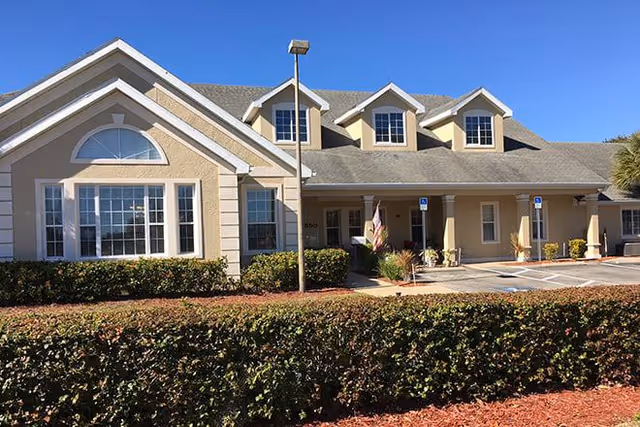 Exterior front view of a single-story building with beige walls, white trim, multiple windows, and a gabled roof under a clear blue sky. There is a hedge and landscaped area in front, a parking area with handicap parking signs, and a covered entrance.