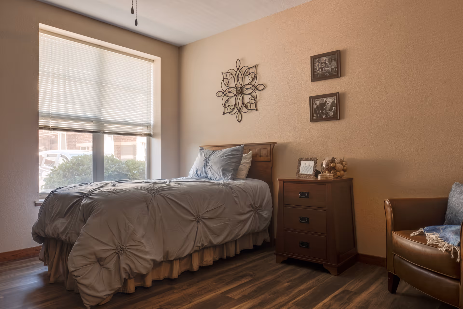A cozy bedroom with a single bed covered in a gray comforter and pillows, a wooden nightstand with decorative items and framed photos, a brown leather armchair with a blue pillow and blanket, and a large window with blinds letting in natural light.