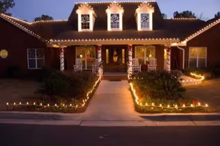 Front entrance of a brick assisted living building lit at dusk with white string lights along the roofline, porch columns, and pathways.