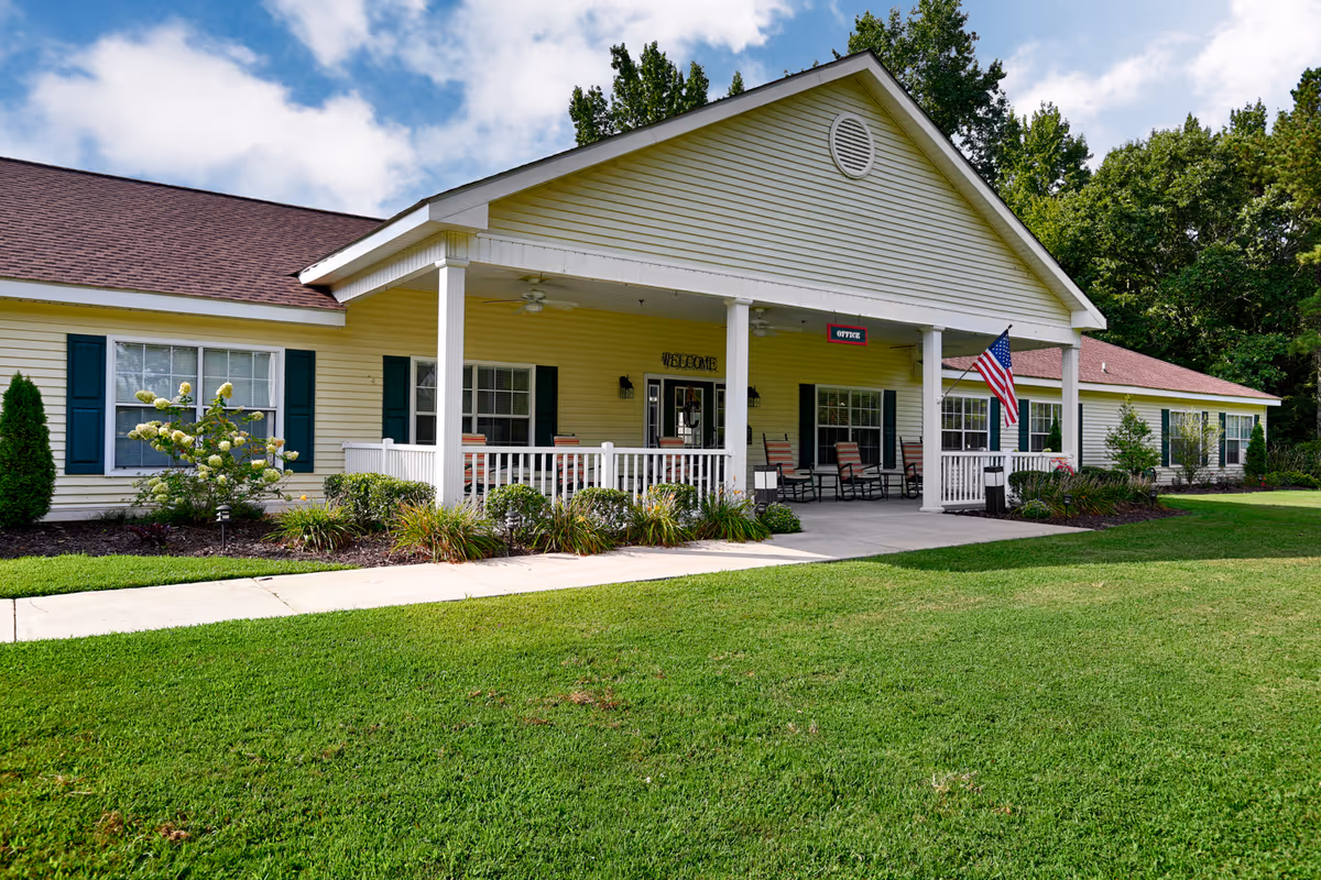 Single-story yellow senior living building with a covered front porch, rocking chairs, and an American flag on a manicured lawn.